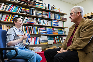 a man and student sitting in chairs facing each other while having a conversation in front of a bookcase
