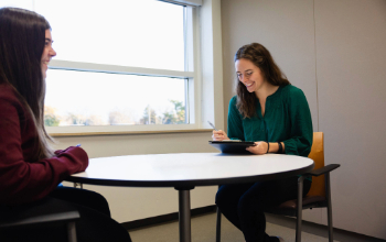 Two women sit across from each other at a round table in a brightly lit room. One woman, holding a pen and notepad, smiles as she listens.