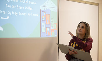 A young woman with long blonde hair and wearing a sweatshirt points to a presentation projected onto a white screen.