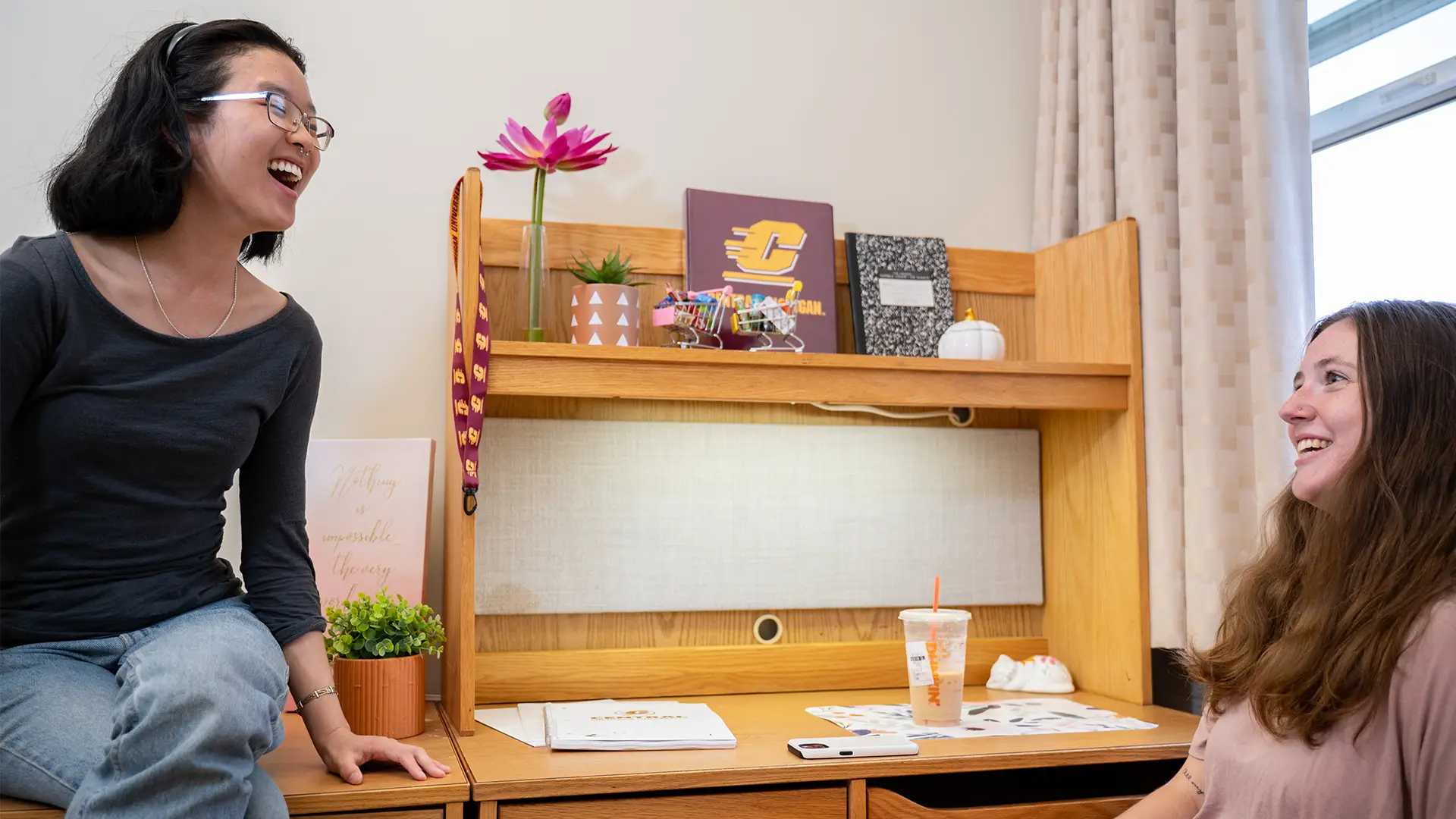 Two students sitting in a dorm room, one is laughing and talking to the other, and a wooden desk is in the background with notebooks, a potted plant, and coffee on the desk.