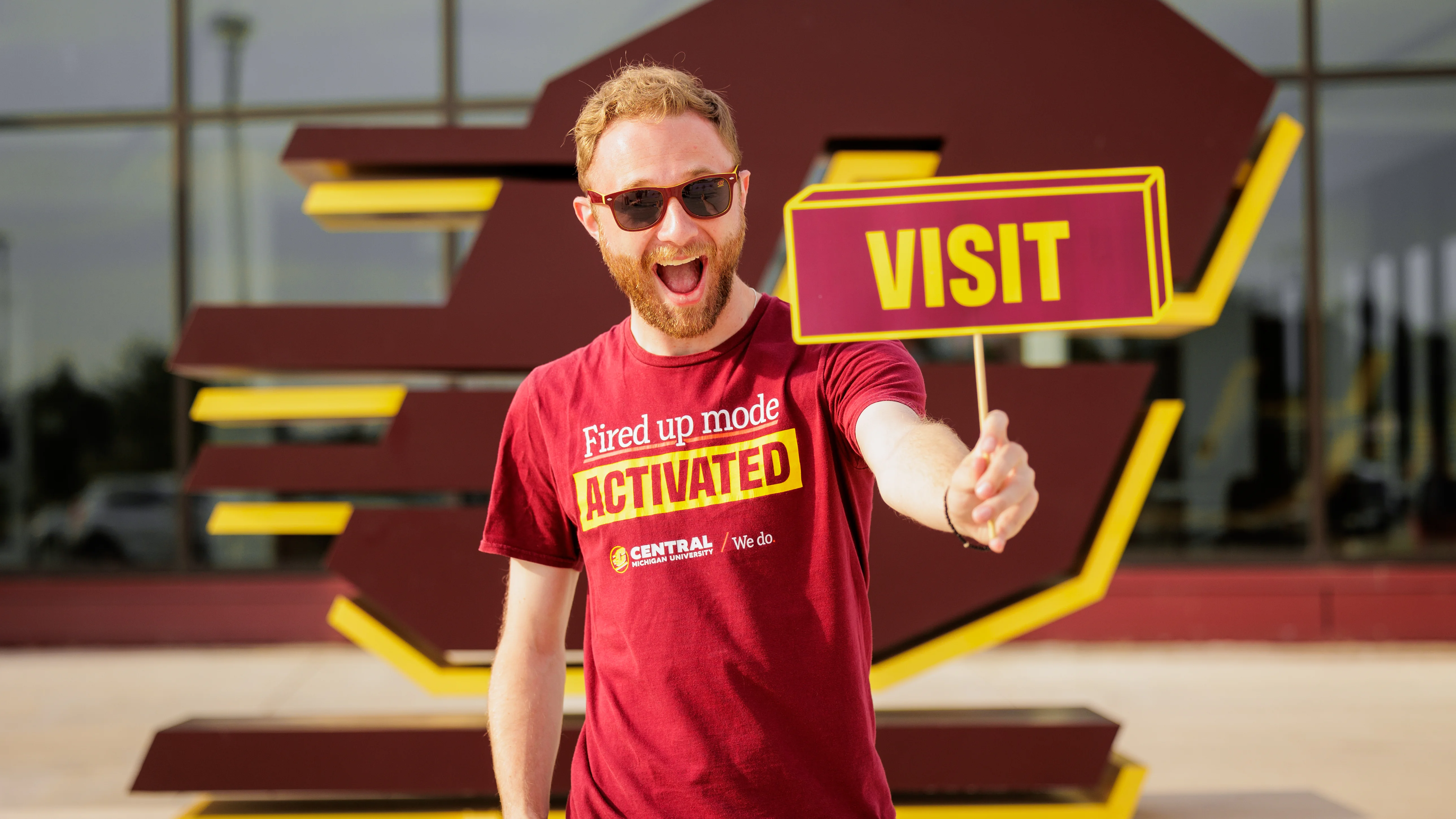 A student wearing a maroon CMU T-shirt that says Fired Up Mode Activated, smiling broadly and wearing sunglasses, holds a maroon and gold sign that says Visit. In the background is a large Action C statue on campus.