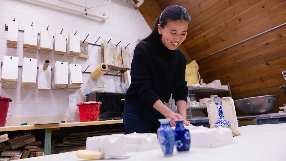 A student smiles as she works on a clay piece in a pottery studio. There are three small blue pots on the table, along with clay and tools.