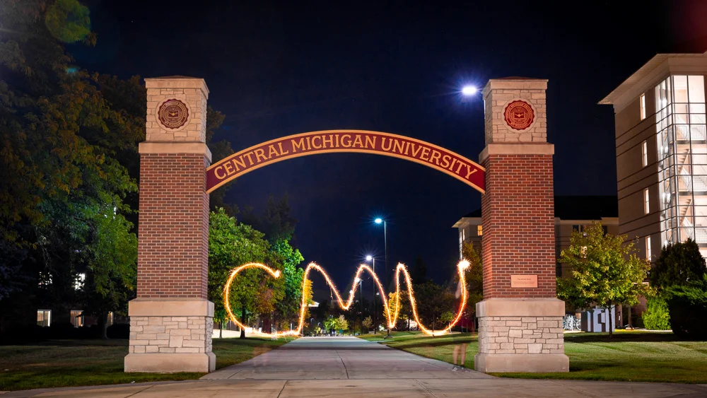 Central Michigan University archway with a dark, blue night sky in the background. The letters CMU are written out in the air with a sparkler and glow yellow with a time lapse.