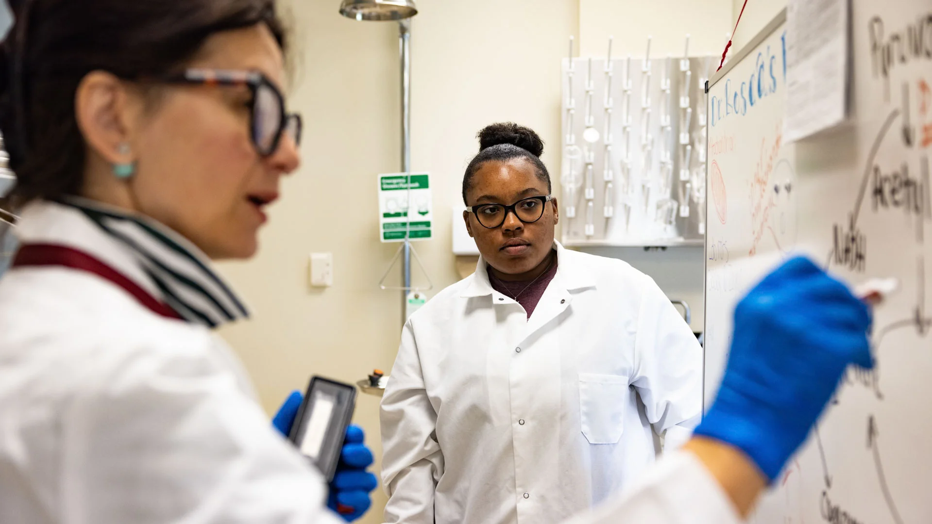 A student in a white coat and glasses looking at a professor in a white coat and blue safety gloves, while they are both inside of a lab.