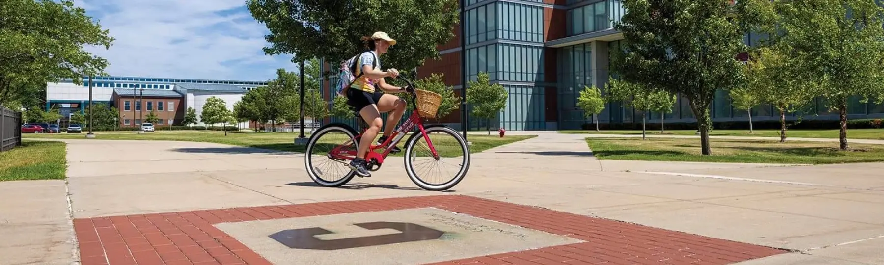 A female rides a red bike across campus in the summer, passing in front of a large bioscience building.