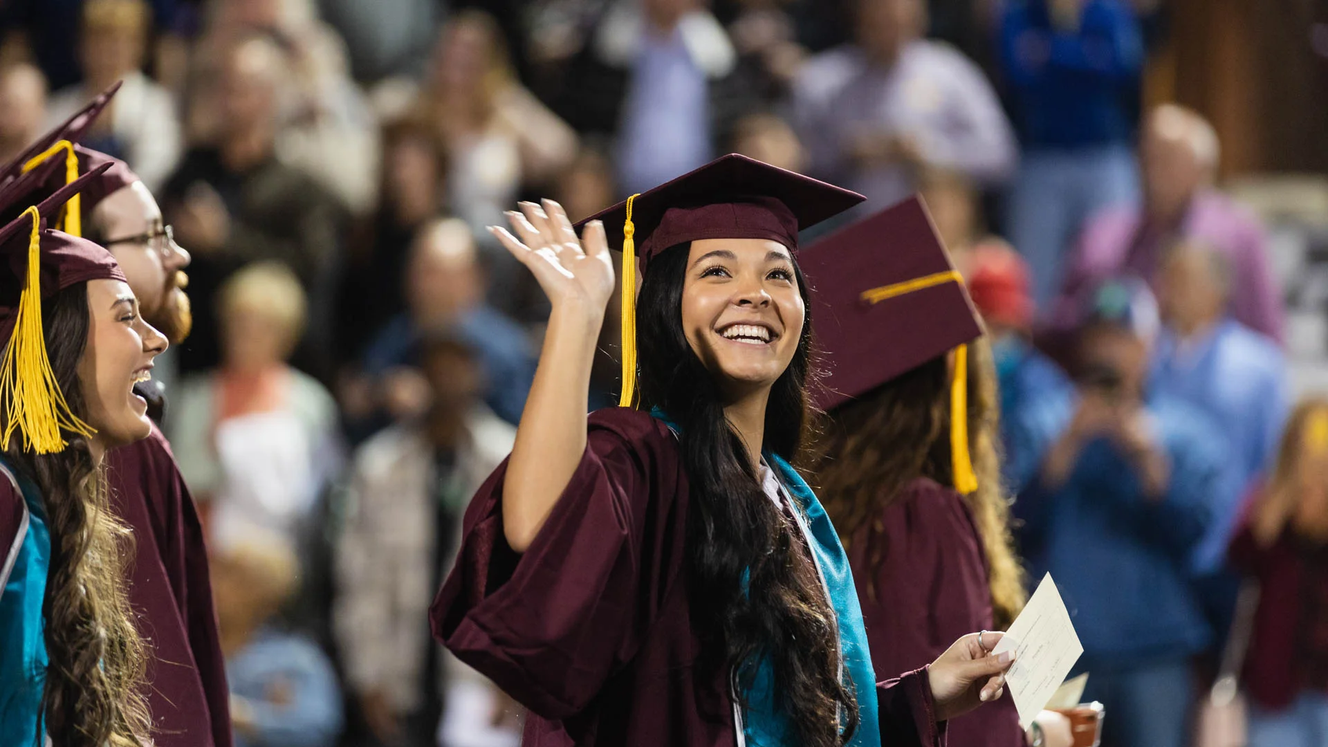 A Central Michigan University student in a maroon graduation gown and cap, smiling and waving during commencement.