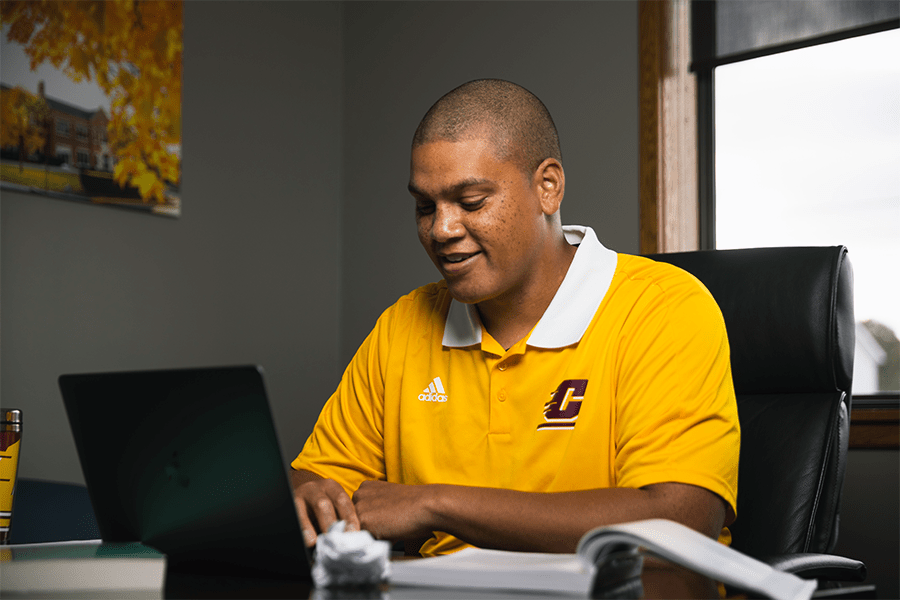 A man in a yellow shirt working on his laptop with a textbook open on a table.