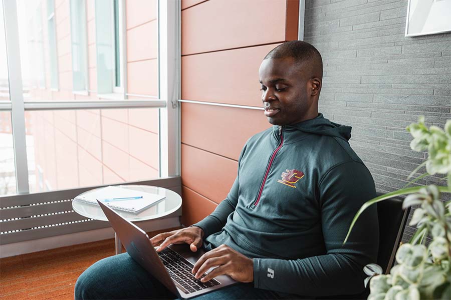 A man sitting near a table with a notebook and pen resting on it as he types on a laptop in a well-lit office