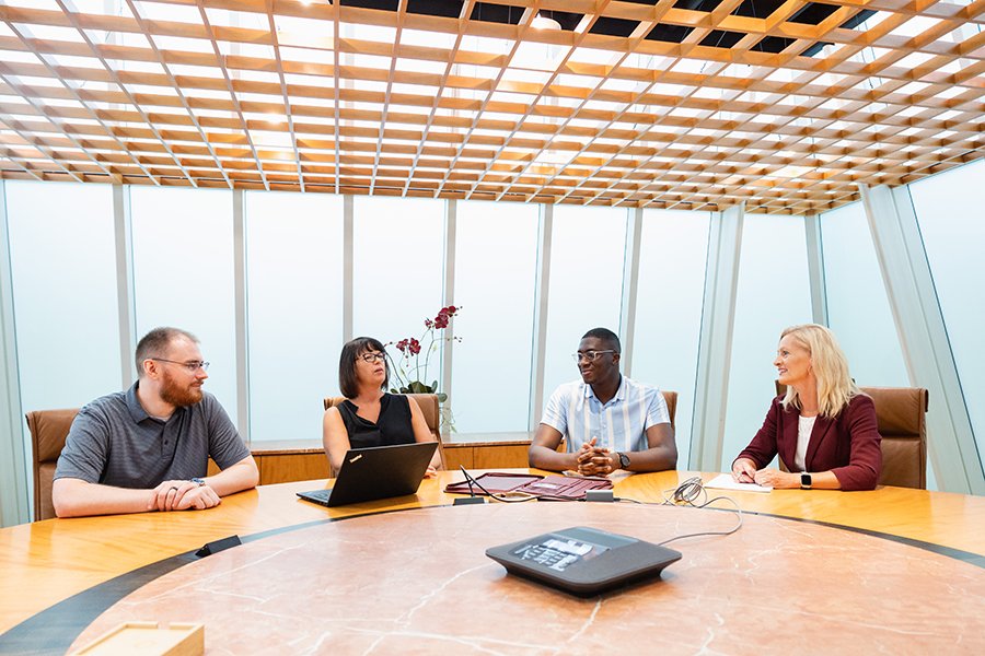 A group of people sitting around a large table having a discussion with an open laptop and folders in front of them.