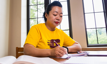 Student in a CMU shirt taking notes in a study space with natural light from tall windows.