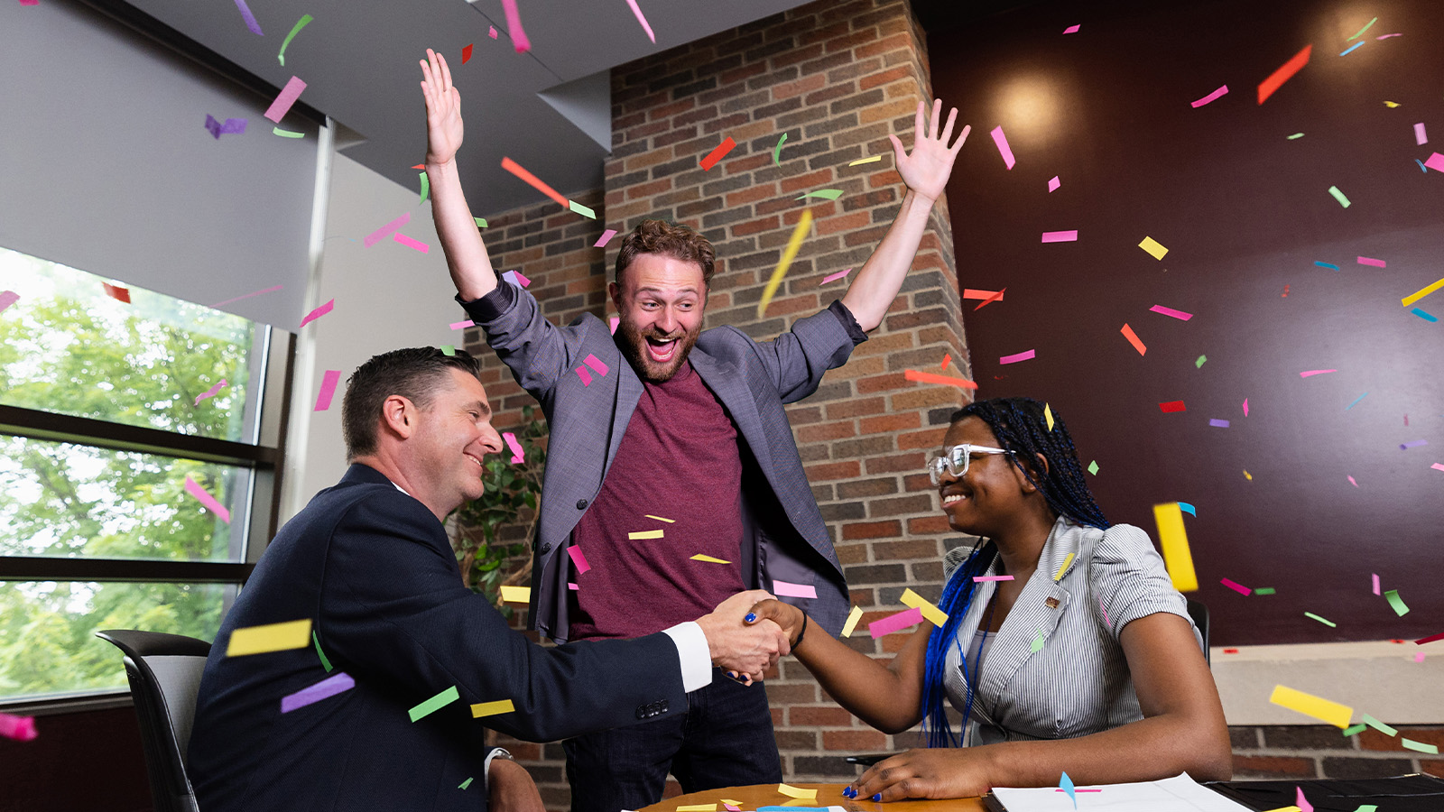 1600x900_Hero_HND_RWR_IMD_fam1 Two people in professional clothes shake hands at a conference table. A person stands in the background cheering in excitement while confetti falls.