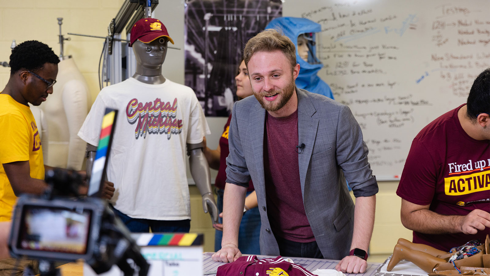 A student stands behind a table, wearing a blazer and maroon shirt, looking at a camera. Surrounding him are other students working on or fitting human mannikins.