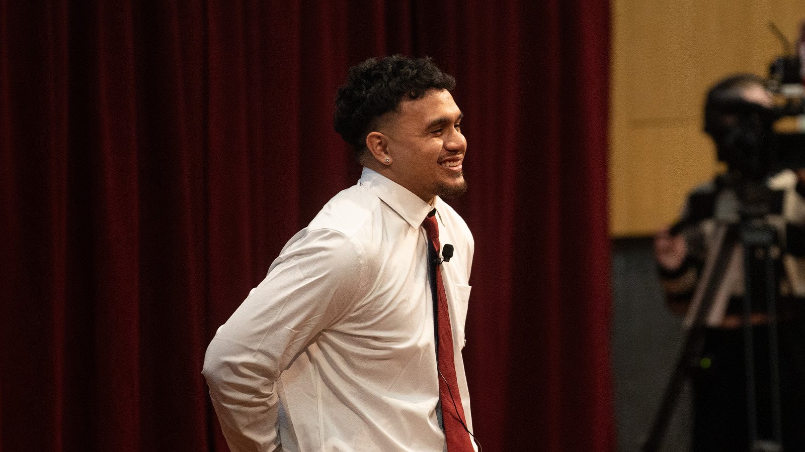 1600x900_Lawai'a Brown Feature A CMU student wearing a dress shirt and tie smiles on stage. A microphone is clipped to his lapel.