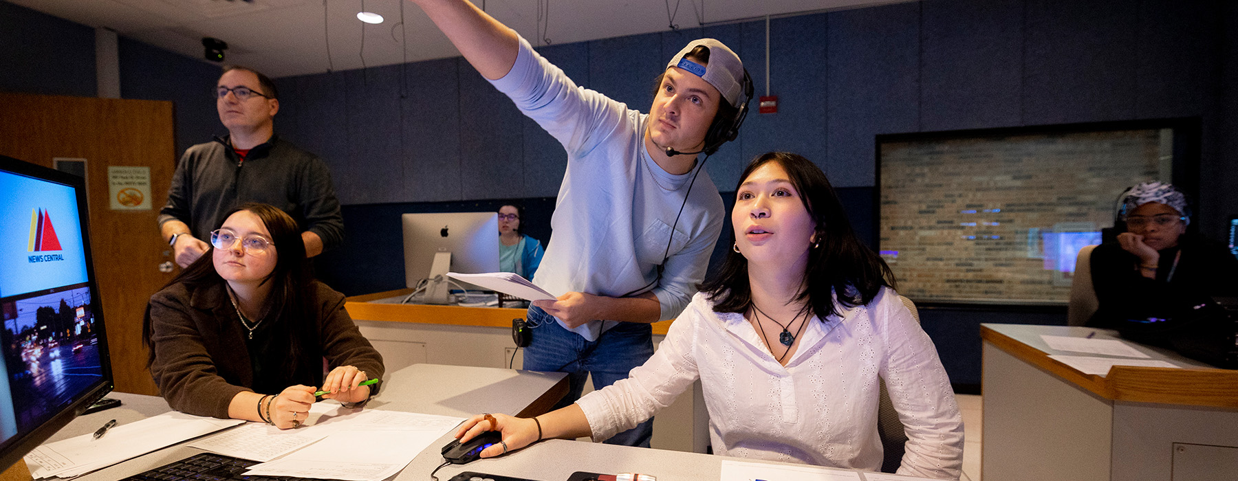 Two students collaborate in a control room of a T.V. studio. One person wears a headset and points at a screen. The other works at computers and technical equipment.