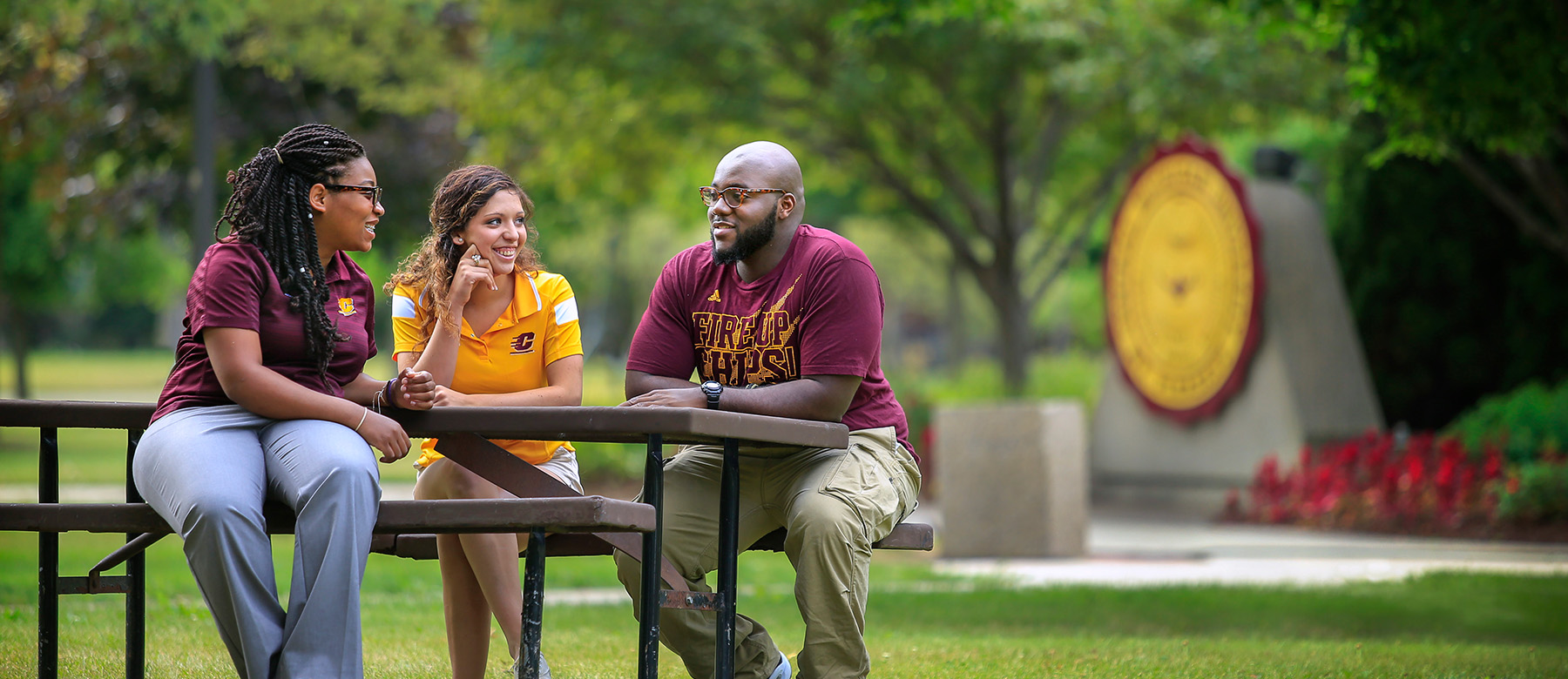 A student sits at a campus picnic bench with two faculty mentors during the summer, with green trees and flowers.