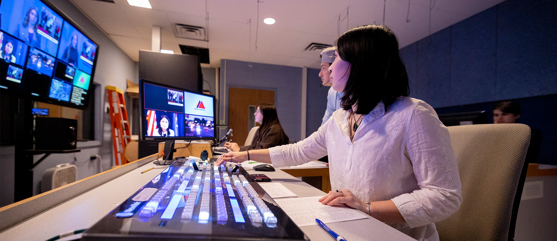 A student sits in a news studio in front of a sound board and a screen displaying multiple camera angles.