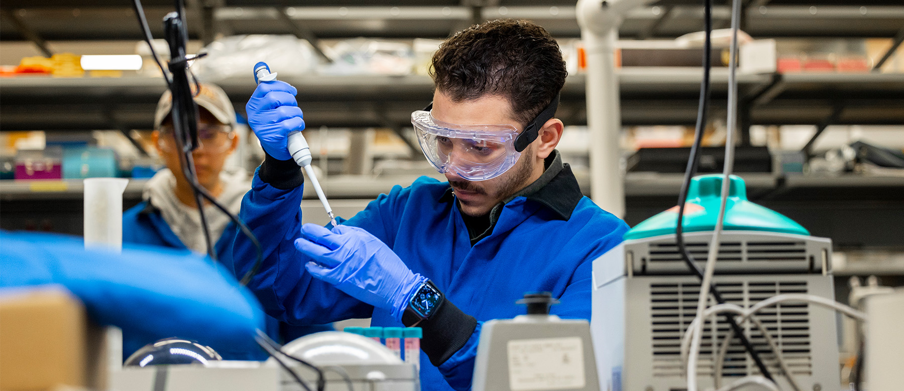 A student sits in a lab wearing a blue lab coat, gloves and safety glasses. The student uses a pipette machine to fill a small vial.