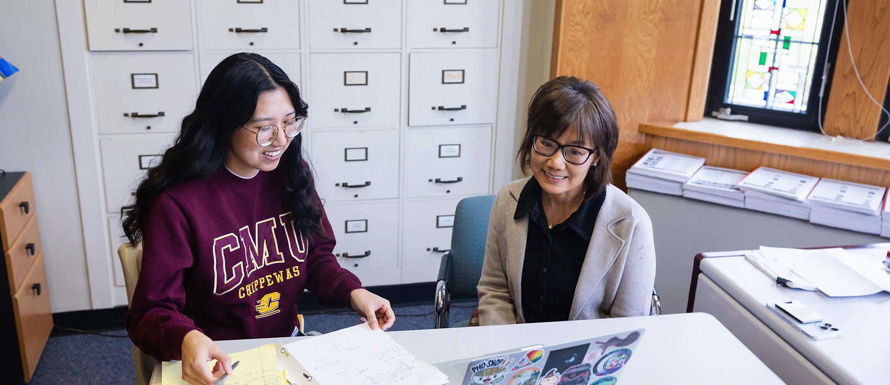A student sitting at a desk with a professor as they look at a computer and papers.