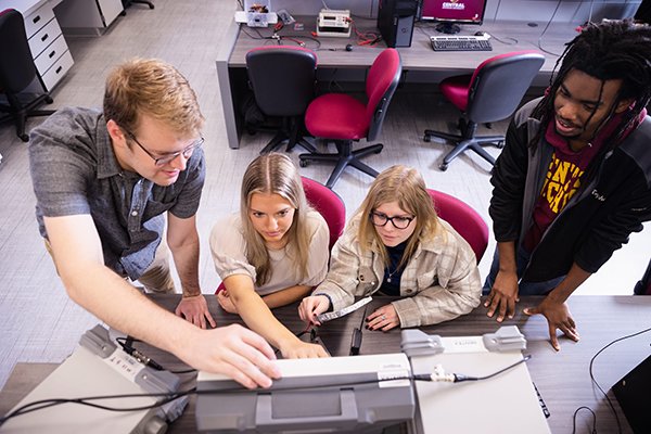 Students in engineering lab gather around a computer pointing at the screen while looking at it focused