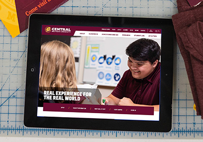 An ipad lay angled across a light gray table, the Central Michigan University home page visible on the screen. Various papers in maroon and gold scattered around the ipad.