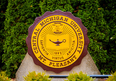 Central Michigan University official seal, a photo of the maroon and gold seal on a smooth stone arch with trees surrounding it outside on CMU’s campus.