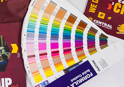 A Pantone swatch book fanned out across a light gray table, an assortment of colors visible, ranging from light pinks to dark brown.