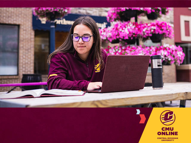 Female student with long brown hair and glasses sits outside on a bench typing on a laptop and reading a textbook. The CMU Online logo sits in the lower right corner, with chevrons pointing to it, on a maroon background.
