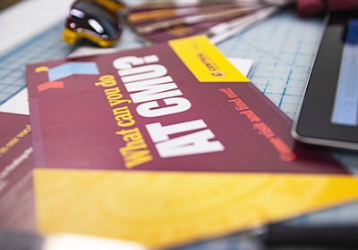 A maroon and gold postcard lays across a table with an ipad, maroon and gold sunglasses next to it, the postcard features red, gold and blue chevrons with the CMU logo in the corner.