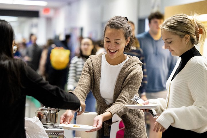 Two students hold paper plates while receiving spoonfuls of international food.