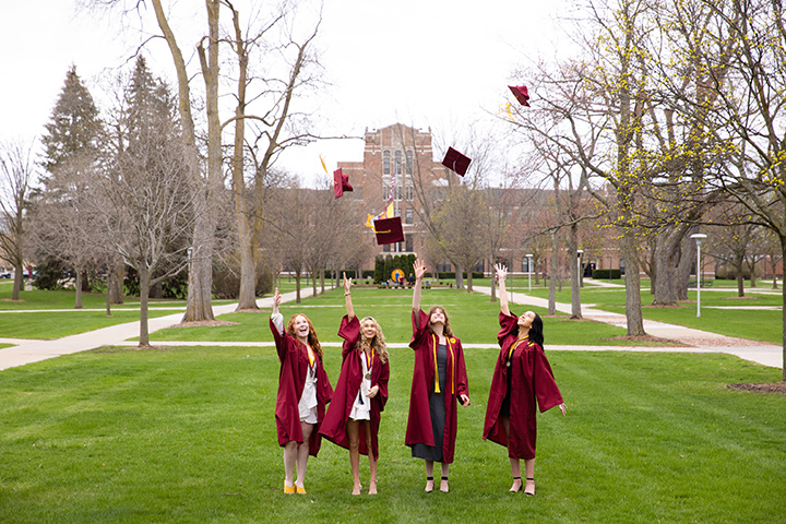 Four CMU students in graduation outfits toss their caps in the air in front of Warriner Hall.