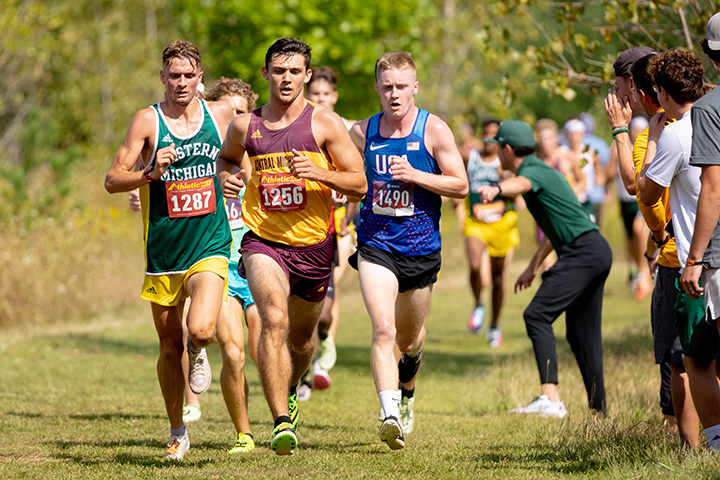 A CMU men's cross country runner leads a group of five runners while spectators cheer.