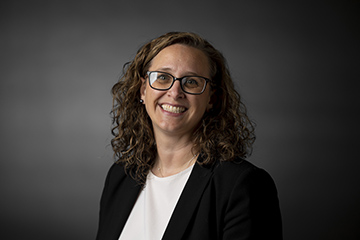 Erica Johnson wears a white shirt and black jacket, smiling at the camera in front of a gray background.