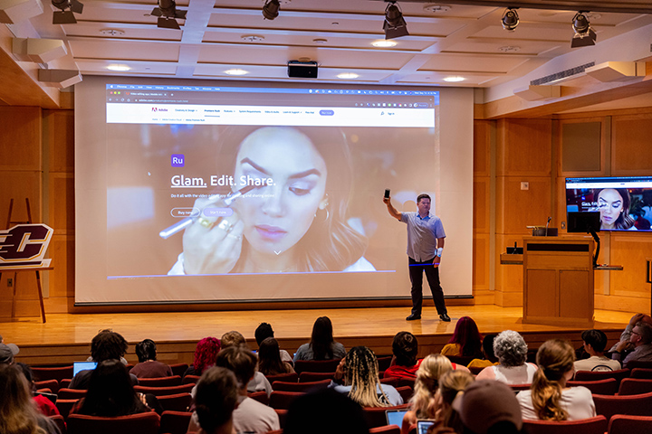 Students, faculty and staff sit an an auditorium while a keynote speaker stands on stage in front of a large projector screen.