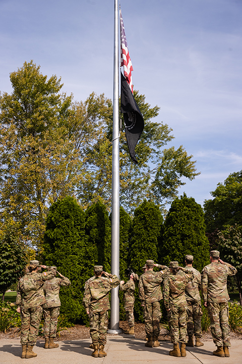 The US and POW flags fly on a flagpole while members of the CMU ROTC program stand nearby at attention.