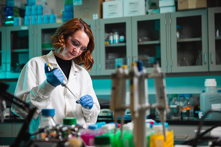 A student wearing a white lab coat and blue gloves works in a lab.