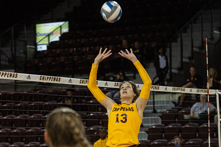 A CMU women's volleyball player wearing a yellow uniform sets the ball near the net.
