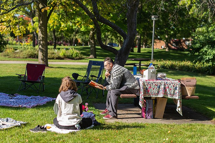 One person sits on a picnic table while another sits on a blanket, both reading poetry.