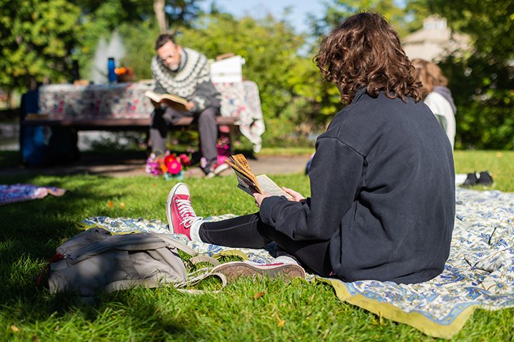 One person sits on a picnic table while another sits on a blanket, both reading poetry.