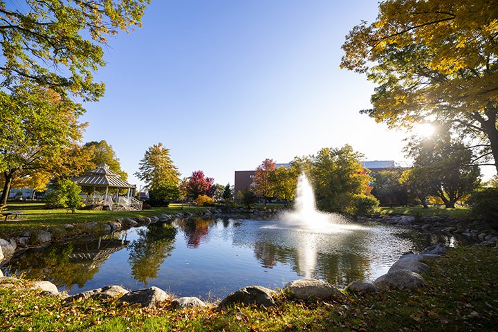 The skies and trees reflect in a pond while a fountain shoots water into the sky.