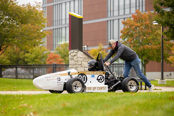 A student pushes a formula race car while another student sits in the driver seat.