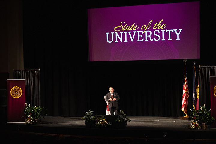 A wide angle shot of CMU President Bob Davies on stage in Plachta Auditorium with a screen behind him with the words 