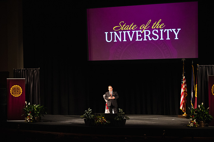 A wide angle shot of CMU President Bob Davies on stage in Plachta Auditorium with a screen behind him with the words