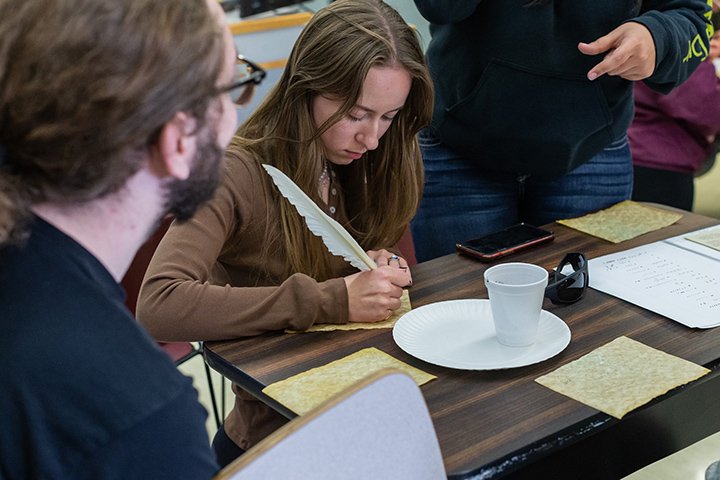 A student writes with a feather quill pen while a professor watches.