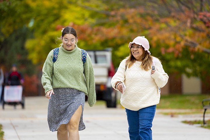 A student in a green sweater and skirt walks to a student wearing a white hat, white sweater and blue jeans.