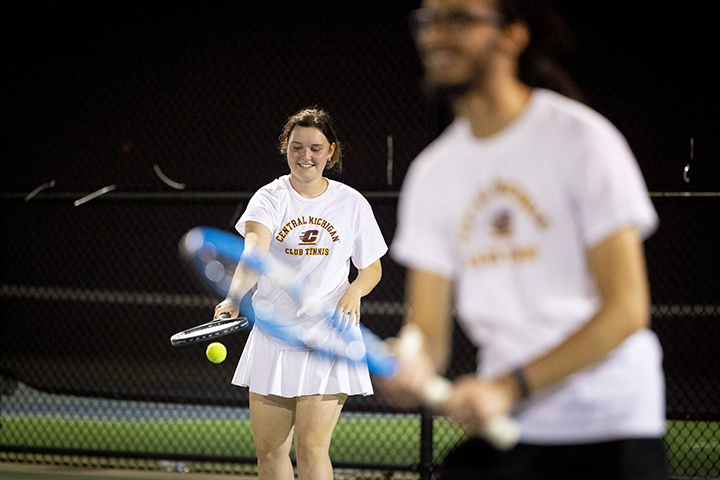 A female student in a white shirt and white skirt holds a tennis racket while bouncing a ball with a blurry male student in the foreground.