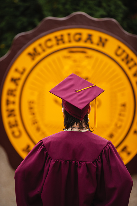 A view from the back of a student in a maroon cap and gown in front of the CMU seal.