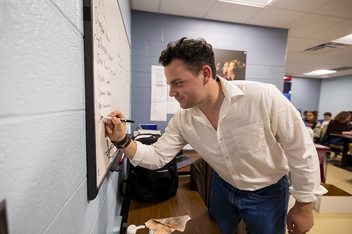 A student in a while collared shirt and jeans write on a white board.