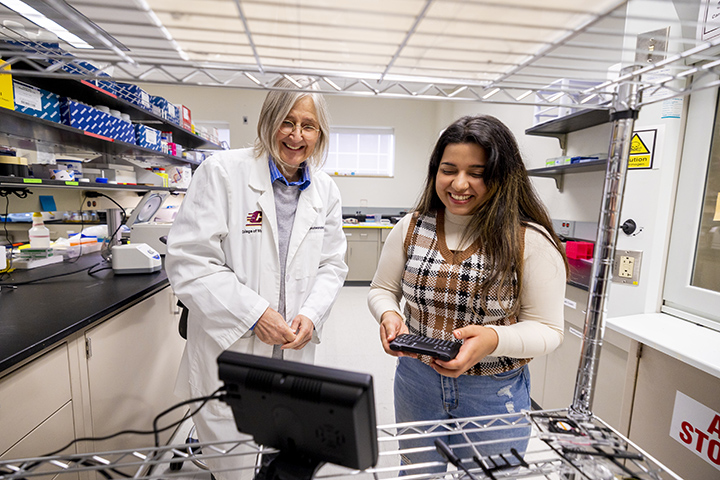 A female student in a plaid shirt stands next to a faculty member in a lab coat while looking at a computer screen.