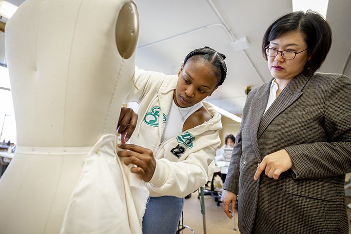 A fashion merchandising student works on a white, cloth mannequin while her professor looks on.