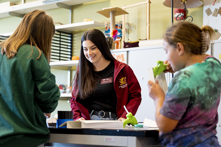 A female student in a maroon CMU shirt stands at a table while working two two fifth grade students.